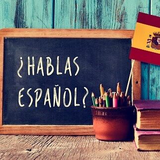 a chalkboard with the question hablas espanol? do you speak Spanish? written in Spanish, a pot with pencils and the flag of Spain, on a wooden desk